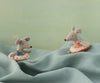 Two Maileg Beach Mouse With Float, Little Sister - Flower toys in swimsuits and colorful float rings sit playfully on wavy blue fabric, with a soft green background behind them.