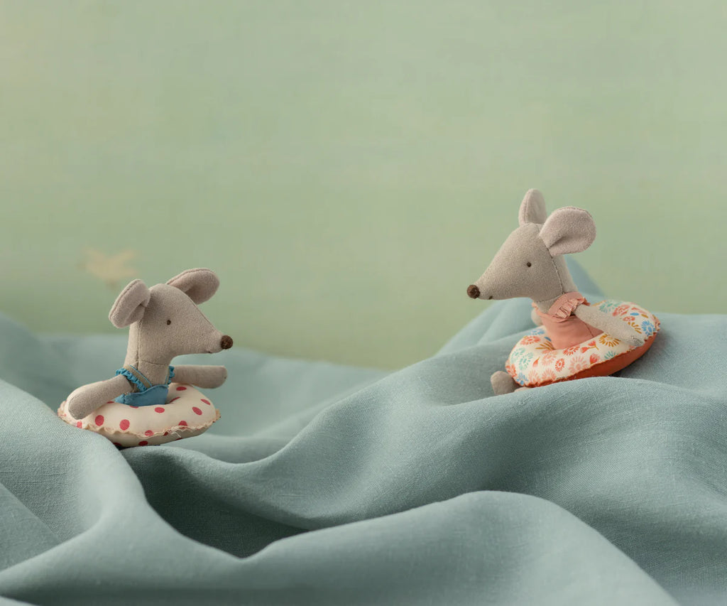 Two Maileg Beach Mouse With Float, Little Sister - Flower toys in swimsuits and colorful float rings sit playfully on wavy blue fabric, with a soft green background behind them.