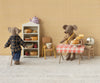 Dad Mouse, dressed in Maileg's extra clothing set—a brown checked shirt and pants—enjoys a cozy miniature kitchen with shelves of bread and jars, while pastries sit on a red-checked tablecloth beside a teapot and covered dish.