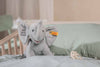 The “My First Steiff” Ellie Elephant Plush Baby Toy, 9 Inches, in gray with "My First Steiff" on its ear, sits on a mint green pillow in a crib—an adorable baby gift. A pacifier lies nearby on the bedding.