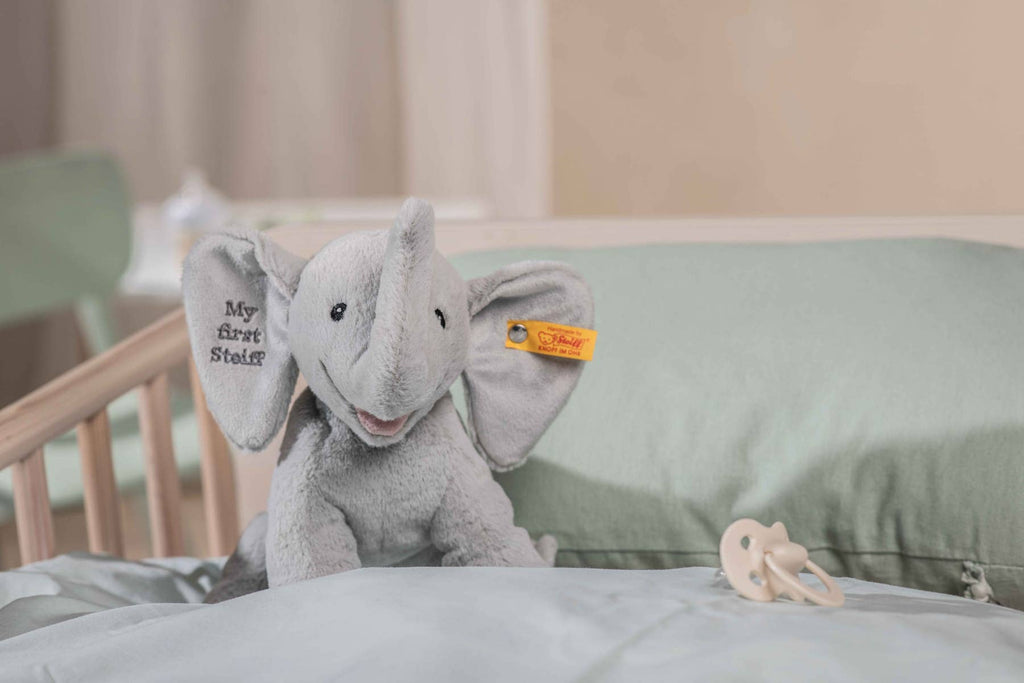 The “My First Steiff” Ellie Elephant Plush Baby Toy, 9 Inches, in gray with "My First Steiff" on its ear, sits on a mint green pillow in a crib—an adorable baby gift. A pacifier lies nearby on the bedding.
