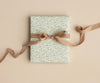A rectangular gift wrapped in pale floral paper, tied with Maileg Ribbon - Soft Brown (10m) in a bow, sits on a beige background.