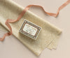 A matchbox labeled "Baby Twins Safety Matches" with illustrations of two babies rests on patterned beige cloth beside Maileg Ribbon - Peach Rose (10m) and pink satin ribbon, creating an exclusive look on a light background.
