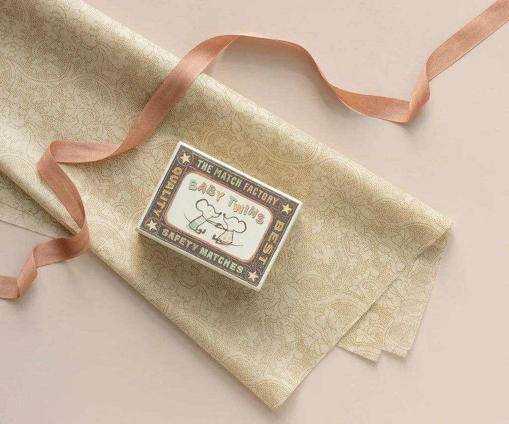 A matchbox labeled "Baby Twins Safety Matches" with illustrations of two babies rests on patterned beige cloth beside Maileg Ribbon - Peach Rose (10m) and pink satin ribbon, creating an exclusive look on a light background.