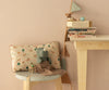 A plush toy rhino sits on a gray stool next to a Maileg Cushion - Unicorn, a miniature chair, and a blanket. Nearby, a wooden table displays stacked books, a toy teapot, and printed fabrics draped as garland.
