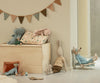 A cozy children's room features soft toys, a wooden toy chest, small furniture, a Maileg Cushion - Unicorn made from printed cotton fabrics, and a string of triangular flags on a beige wall.