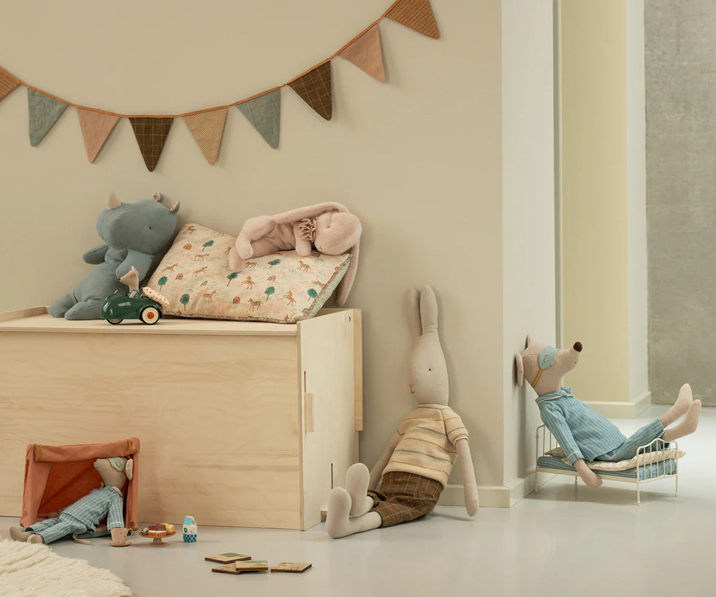 A cozy children's room features soft toys, a wooden toy chest, small furniture, a Maileg Cushion - Unicorn made from printed cotton fabrics, and a string of triangular flags on a beige wall.