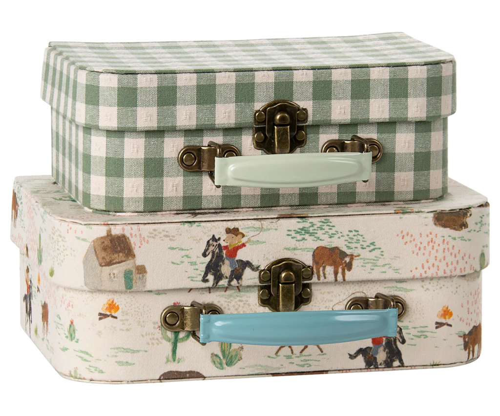 Two small decorative suitcases from the Maileg Cowboy Set are stacked: the top has a green and white gingham pattern with a mint handle, and the bottom shows a farm and cowboy print with a blue handle. Both feature metal clasps.