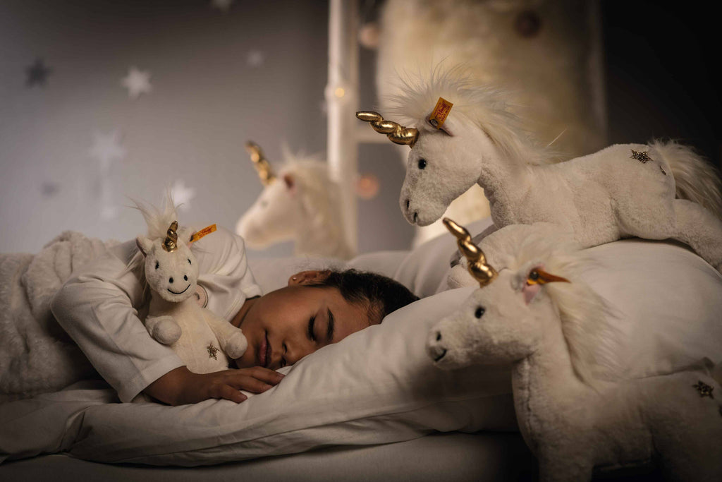 A child sleeps peacefully in bed, cuddling a Steiff Unica Unicorn Stuffed Plush Toy (11 Inches), while other golden-horned unicorn plush toys surround her and stars shine on the wall, creating a magical atmosphere.