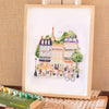 The Paint By Number Kit - Paris features a framed scene of a lively Parisian street with the Eiffel Tower, trees, café, and people. Includes paint pots and brushes beside the frame—perfect for aspiring artists.