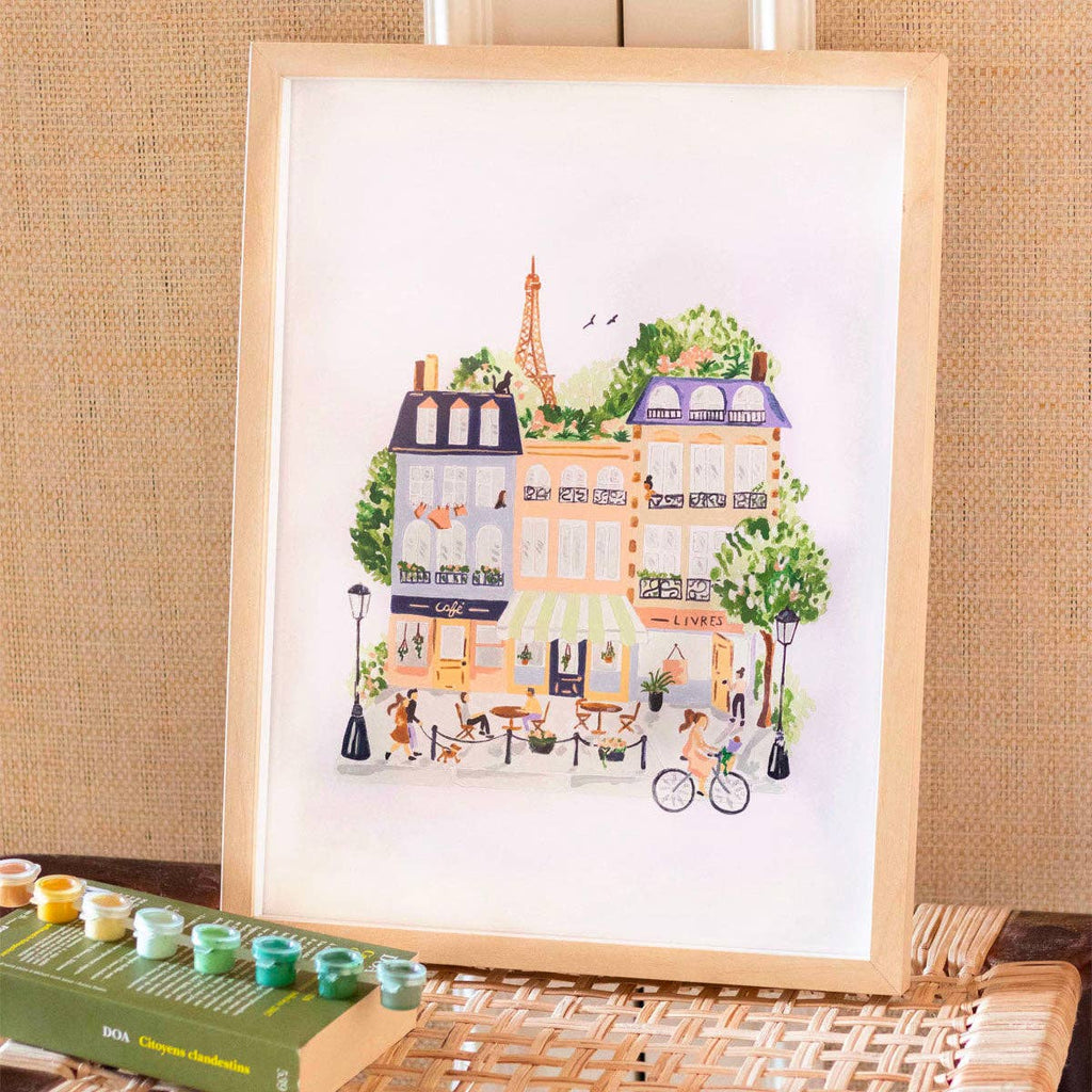 The Paint By Number Kit - Paris features a framed scene of a lively Parisian street with the Eiffel Tower, trees, café, and people. Includes paint pots and brushes beside the frame—perfect for aspiring artists.