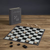 The Board Game: Checkers & Backgammon Vintage Bookshelf Edition features a black-and-white board with black and beige pieces, presented in a closed, upright book-style case on a wooden table—perfect for fans of classic games.