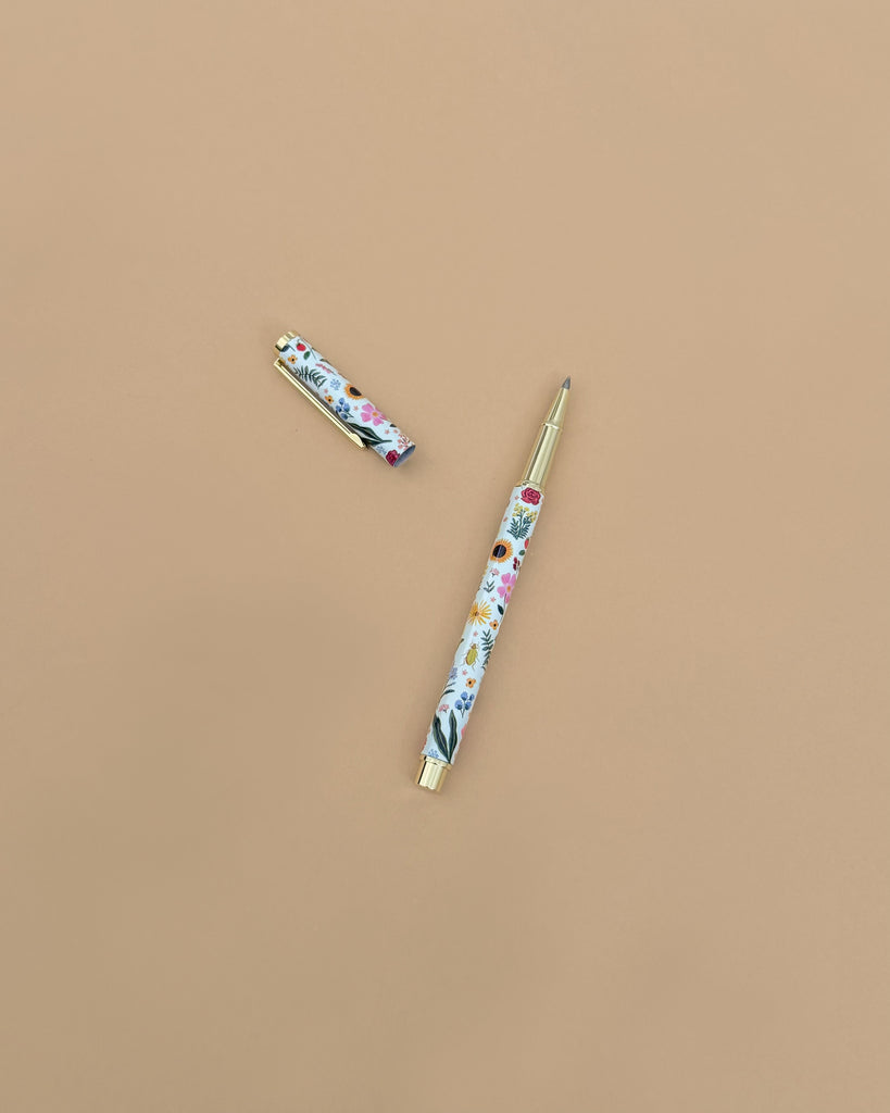 The Curio Pen, featuring a floral pattern with a gold clip and tip, rests on a light brown surface with its cap nearby. It’s compatible with gel ink refills for smooth writing.