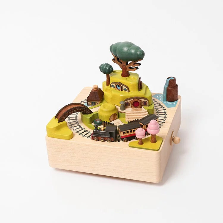 A Wooden Train Music Box - Spring from Wooderful Life featuring a train, tracks, trees, a tunnel, and two figurines on a platform. The structure has a handle, suggesting manual motion activation.