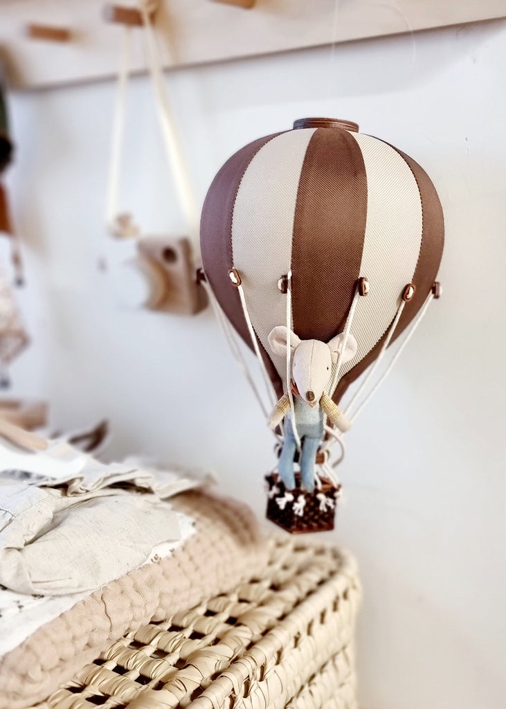 The Hot Air Balloon - Light Brown / Dark Brown ornament, featuring a small stuffed mouse toy, hangs above a woven basket and adds charm to a cozy, softly lit children’s room.