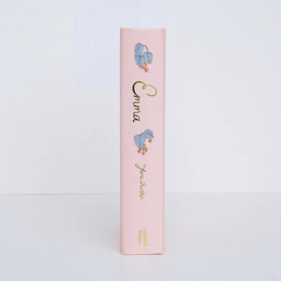 The spine of the light pink hardcover "Emma | Collector's Edition" stands upright, featuring gold foil lettering and blue flower illustrations, set against a white background.
