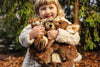 A young child in a white, fluffy coat smiles while holding three stuffed toys outdoors, including the Steiff Joggi Hedgehog Plush Toy (6 Inches), with green trees and fallen leaves in the background.