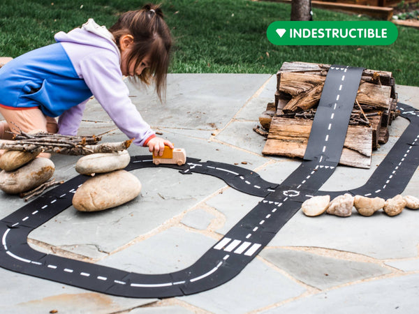 A young child enjoys creative play outdoors on a stone patio with the Large Flexible Toy Road Set - Highway (24-Piece) and a toy car. The child-safe, indestructible black road winds among rocks and firewood stacks.