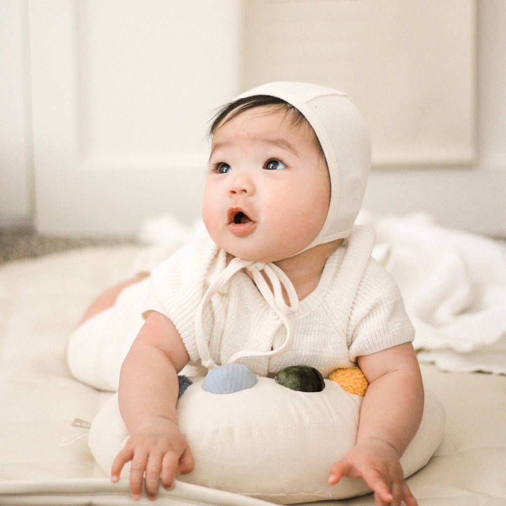 A baby in a cream outfit lies on their stomach, gazing upward with curiosity, hands resting on the Tummy Time Palette—a tummy time pillow featuring colorful patches—while on a soft mat.