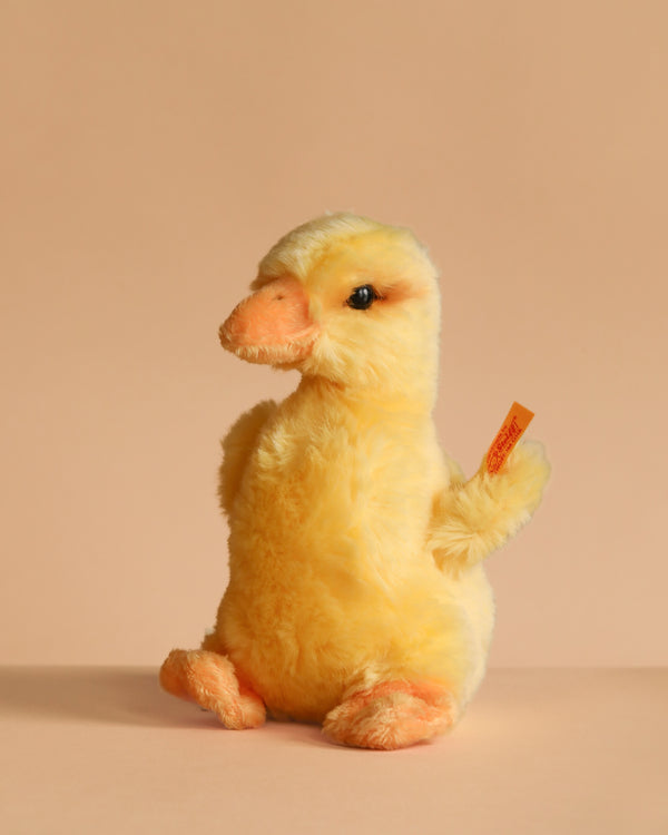 The Steiff Pilla Duckling Stuffed Animal is a soft, yellow plush toy with an orange beak, feet, and a small orange tag on one wing. It sits upright against a plain beige background.