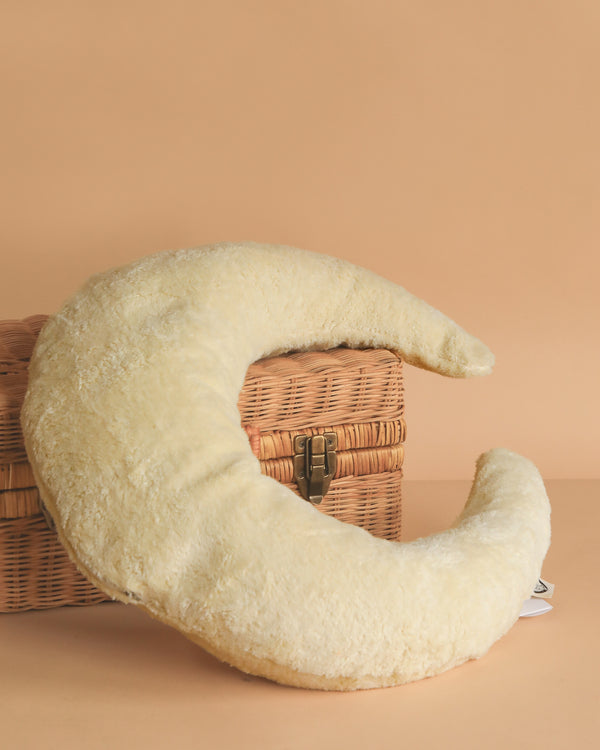 The Senger Naturwelt - Moon Nursing and Neck Pillow, plush and crescent-shaped, rests against a closed wicker basket on a beige background.