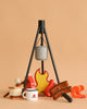 The Wooden Camp Fire Set includes a gray pot on a black tripod, toy food (bread, marshmallows, sausages), and a white mug with a red campfire logo—ideal for kids’ pretend indoor camping adventures.