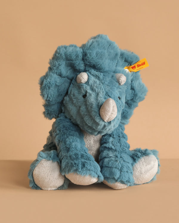 An 11-inch Steiff Dixi Triceratops Dinosaur Plush Toy sits on a beige background, featuring the signature yellow Steiff tag on its left ear that guarantees its authenticity and premium quality.