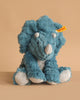An 11-inch Steiff Dixi Triceratops Dinosaur Plush Toy sits on a beige background, featuring the signature yellow Steiff tag on its left ear that guarantees its authenticity and premium quality.