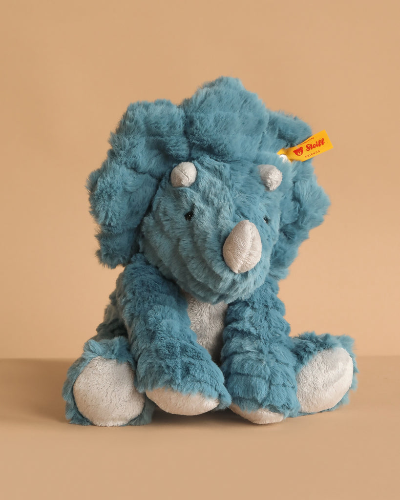 An 11-inch Steiff Dixi Triceratops Dinosaur Plush Toy sits on a beige background, featuring the signature yellow Steiff tag on its left ear that guarantees its authenticity and premium quality.