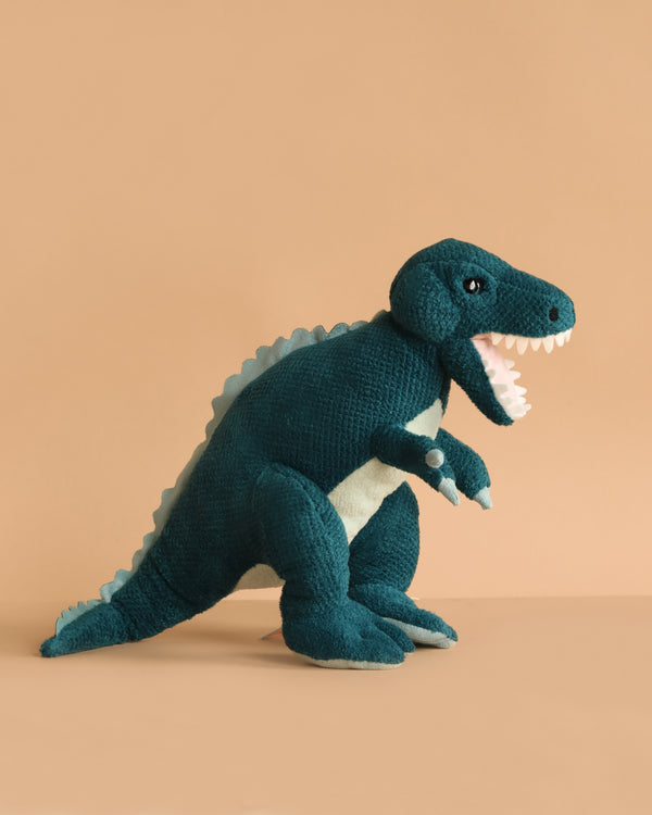 The Steiff Thaisen Tyrannosaurus "T-Rex" Dinosaur Plush Toy, 14 Inches, in dark teal with white felt teeth and a light blue spine, sits on a peach background facing right with its mouth open.