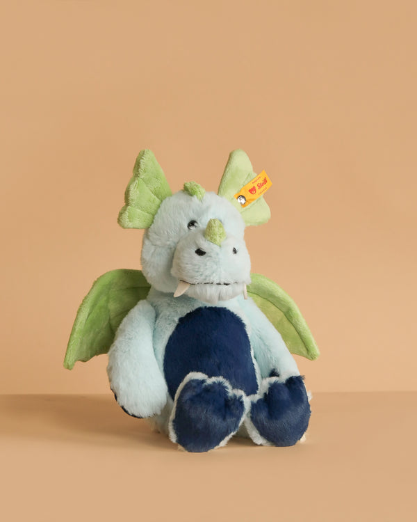 The Steiff Samu The Magic Dragon Plush Toy (11 inches) features light blue, dark blue, and green fur, wings, two tusks, and a yellow ear tag—perfect for kids. It sits against a plain beige background.