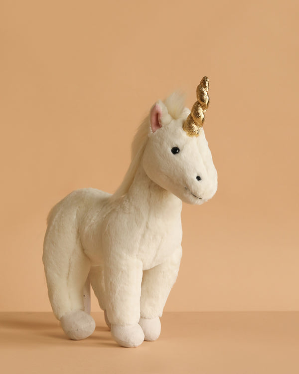 The Steiff Unica Unicorn Stuffed Plush Toy, 11 Inches, stands on a beige surface with a matching background. This plush features a golden horn, black eyes, a fluffy mane, and a gentle whimsical expression.