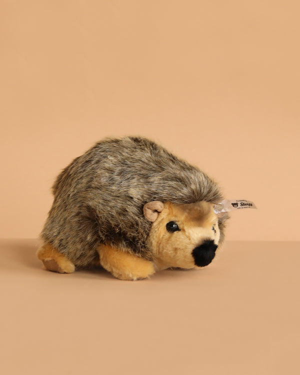 The Steiff Joggi Hedgehog Plush Toy, 6 Inches, features brown and tan fur and sits on a beige surface with a matching background. A small fabric tag on its side adds extra charm to this adorable handmade stuffed animal.