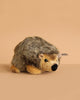The Steiff Joggi Hedgehog Plush Toy, 6 Inches, features brown and tan fur and sits on a beige surface with a matching background. A small fabric tag on its side adds extra charm to this adorable handmade stuffed animal.