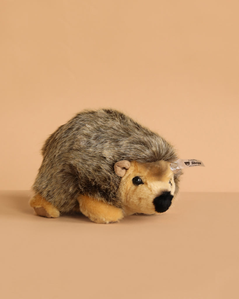 The Steiff Joggi Hedgehog Plush Toy, 6 Inches, features brown and tan fur and sits on a beige surface with a matching background. A small fabric tag on its side adds extra charm to this adorable handmade stuffed animal.