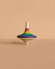 The Mader Large Spinning Top - Rainbow, featuring vibrant stripes, stands upright on a beige surface against a matching beige background.