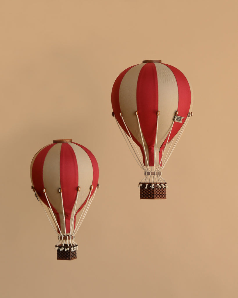 Two burgundy and white striped handmade Hot Air Balloon - Burgundy decorations with wicker baskets float against a beige background. Vintage-inspired, they add whimsical charm to any children's room or space.