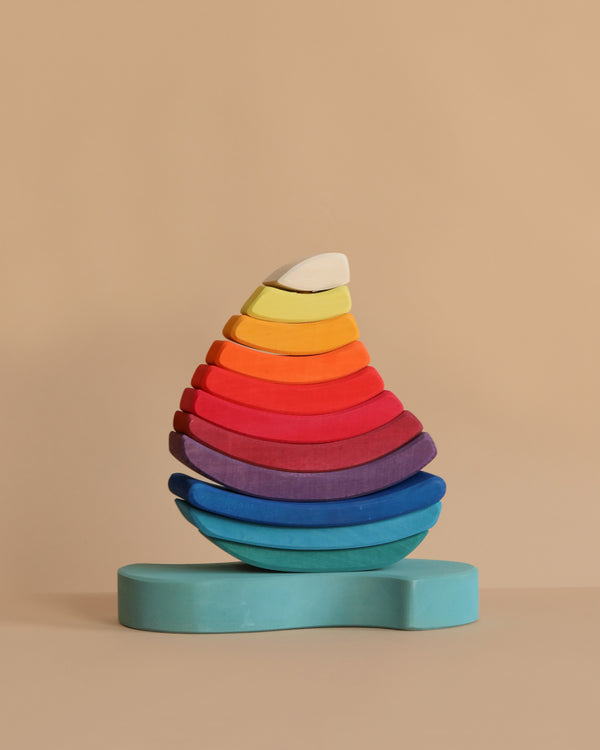Grimm's Stacking Tower Sailboat features semi-circular wooden blocks, arranged by size and color in a rainbow teardrop shape on a light blue base—perfect for fine motor skill development against a beige background.