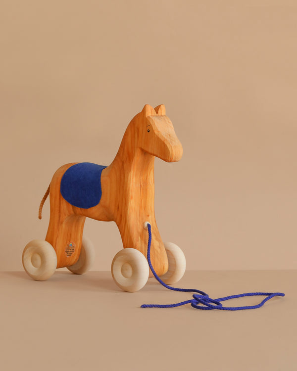Grimm's Horse Holly, a charming wooden pull along toy with white wheels, a blue string, and a blue saddle, rests on a neutral beige surface against a soft beige background.