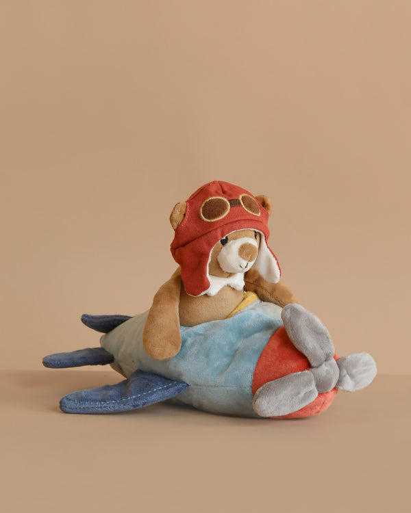 The Pilot Bear Plush Toy features a cuddly bear with a red aviator helmet and goggles, sitting on a soft blue and red vintage airplane, all set against a plain beige background.