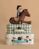 A Maileg Cowboy Set features a cowboy mouse on a plush rocking horse, displayed atop two Maileg suitcases—one green plaid, one with a pastoral print—arranged against a beige background.