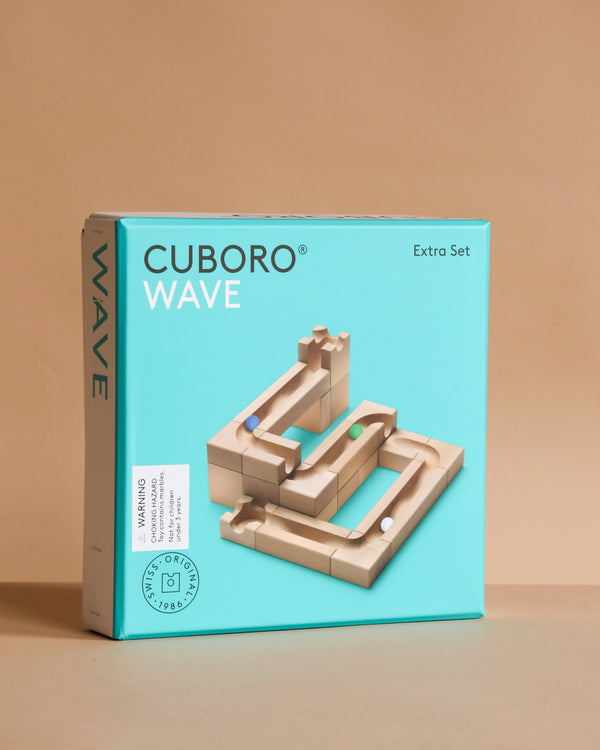 A turquoise box of the Cuboro Wave Marble Run Extra Set, displaying wooden marble run pieces with four marbles arranged on the front. The box stands on a neutral background, highlighting this creative marble run system.