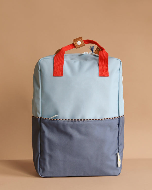 A Sticky Lemon Backpack Large in Bissou Blue + Blue Whale stands upright against a beige background, featuring a color-blocked design, red handles, front pocket, and a small brown tab with white button. Made from recycled PET.