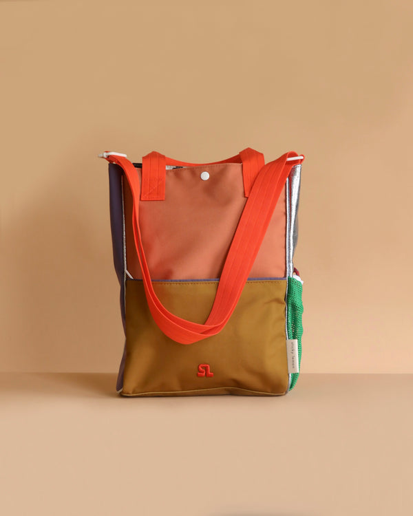 The Sticky Lemon Shoulder Bag | Leather Ball + Golden Cup features orange handles, a brown and tan front panel, green mesh side pocket, small white logo on the front—all crafted from recycled PET bottles—shown against a beige background.