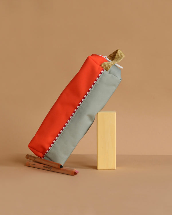 The Sticky Lemon Pencil Case | Color Blocking | Lifebuoy Orange + Shark Grey, made from recycled PET bottles, stands upright next to two colored pencils on a beige background, highlighting its orange and gray color block design.