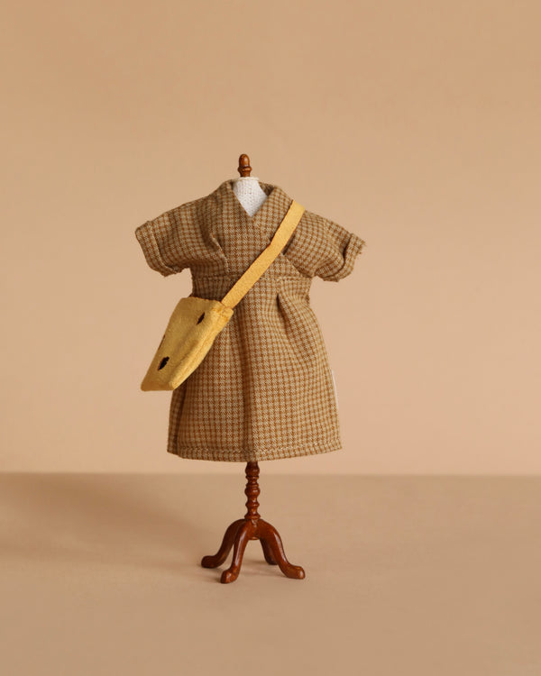 A small mannequin showcases the Maileg Extra Clothing: Dress And Bag For Mom Mouse—a brown plaid short-sleeve dress with a yellow crossbody bag—ideal for doll accessories, set against a plain beige background.