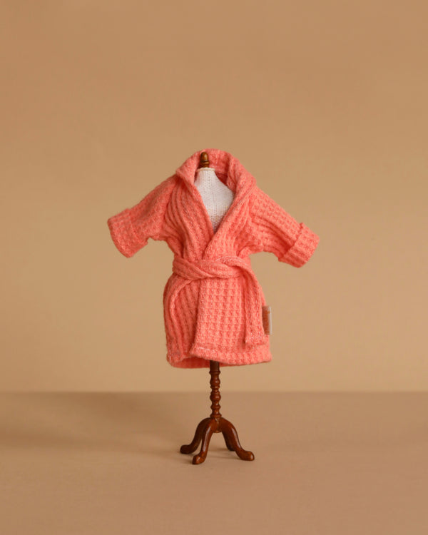 A small mannequin displays the Maileg Extra Clothing: Bathrobe - Coral, a textured pink cotton robe with a belt, set against a plain beige background.