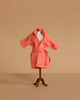 A small mannequin displays the Maileg Extra Clothing: Bathrobe - Coral, a textured pink cotton robe with a belt, set against a plain beige background.