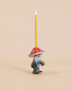 A whimsical Mushroom Gnome Cake Topper, hand-painted and crafted from fine porcelain, with a red and white mushroom cap, holding a lit yellow candle, set against a plain beige background.