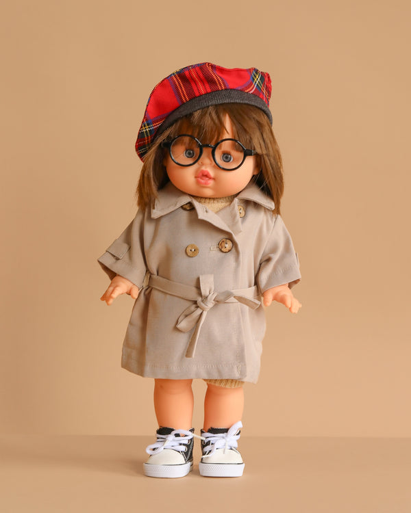 The Minikane Standing Doll (14") - Chléa With Clothing features brown hair, round glasses, and a red plaid beret, dressed in a beige trench coat and white sneakers. Stands against a beige background. Made in Spain.